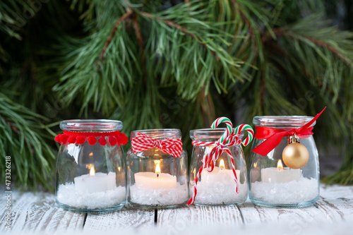 Four candles in glass jars with fir on holiday background. Cozy handmade holiday home decor: glass jar with candle decorated with red ribbon. Christmas decorations.