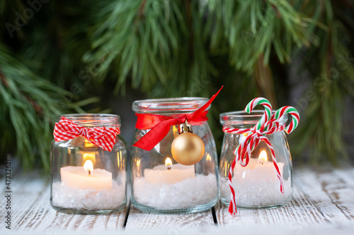 Three candles in glass jars with fir on holiday background. Cozy handmade holiday home decor: glass jar with candle decorated with red ribbon. Christmas decorations.