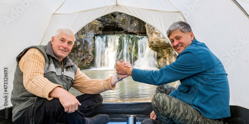 Mature male tourists sit in a tent, shake hands and admire the beautiful Atysh waterfall on a sunny autumn day. Bashkortostan photo