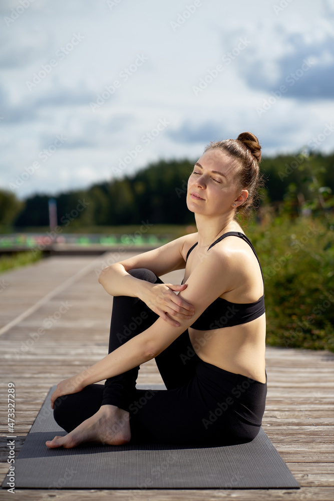 Portrait of attractive young sporty young woman practicing yoga on yoga mat on the wooden pier.
