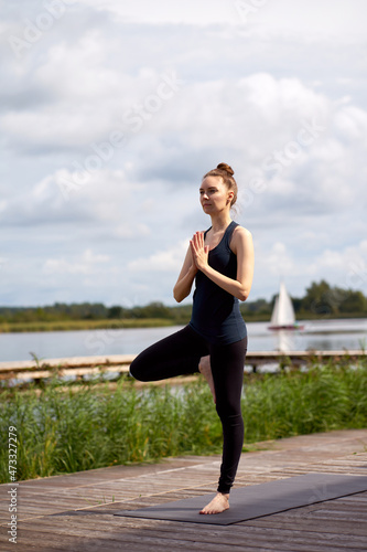 Attractive slim woman wearing black sportswear practicing yoga or pilates near lake.