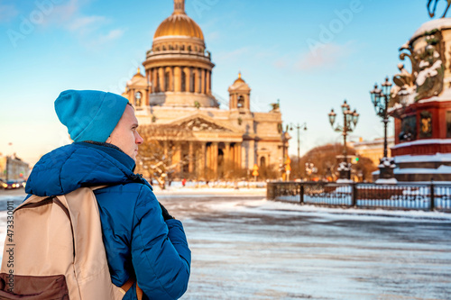 A young man with a backpack stands with a view of St. Isaac's Cathedral and the monument on a snowy winter day in St. Petersburg © KseniaJoyg