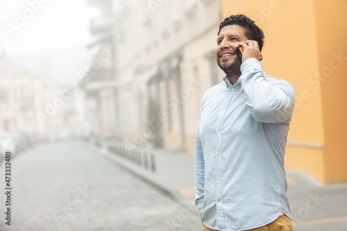 Young cheerful hispanic man talking on phone on foggy european street