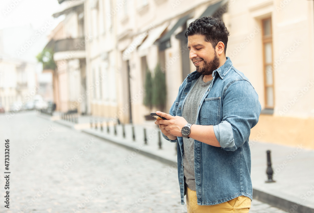Young cheerful hispanic man in jeans jacket using phone on european street Young cheerful hispanic man in jeans jacket using phone on european street