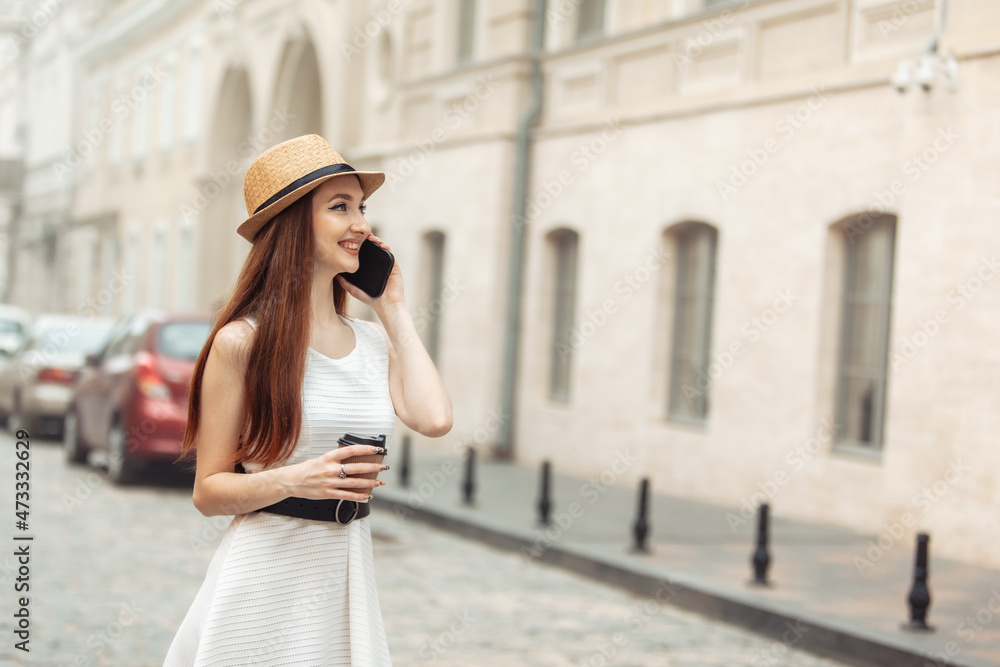 Fototapeta premium Cute caucasian woman in hat talking on the phone and drinking coffee in the city