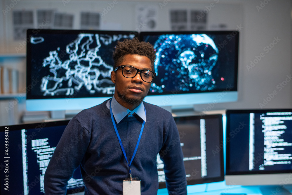 Fototapeta premium Contemporary young programmer standing by workplace against group of computer monitors with coded data on screens