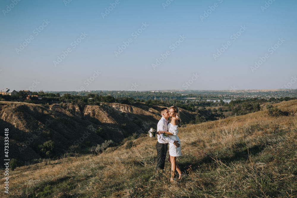 A loving groom and a beautiful blonde bride in a white dress tenderly hug in nature on the background of hills and cliffs in autumn in sunny weather. Wedding photo of smiling newlyweds.
