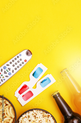 Movie time. Snack bowls, beer bottles, tv remote, 3d glasses on yellow background. Top view. Copy space