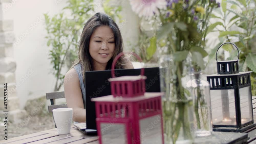 Pretty woman sitting at table in outdoor cafe with laptop. Slider shot of beautiful girl spending time at cafe with beautiful plants, drinking coffee, having video call with friend. Technology concept Pretty woman sitting at table in outdoor cafe with laptop. Slider shot of beautiful girl spending time at cafe with beautiful plants, drinking coffee, having video call with friend. Technology concept