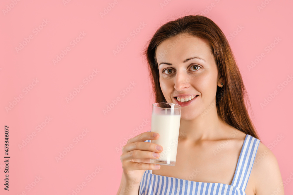 A smiling middle-aged brunette girl drinks milk from a glass glass. Studio portrait close-up on pink background A smiling middle-aged brunette girl drinks milk from a glass glass. Studio portrait close-up on pink background