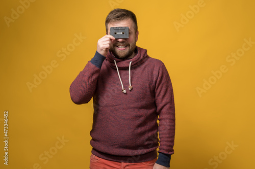 Caucasian bearded man holds an old audio cassette in front of his eyes like glasses and smiles amusingly. Yellow background.