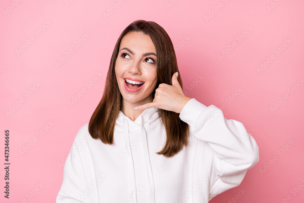 Photo of impressed cute young lady wear white hoodie smiling showing phone gesture looking empty space isolated pink color background Photo of impressed cute young lady wear white hoodie smiling showing phone gesture looking empty space isolated pink color background