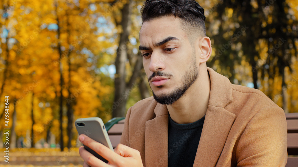 Portrait young serious guy use app hispanic man looking at phone screen browses news on telephone concentrated male sits on bench in autumn park read using device businessman holds smartphone outdoors Portrait young serious guy use app hispanic man looking at phone screen browses news on telephone concentrated male sits on bench in autumn park read using device businessman holds smartphone outdoors