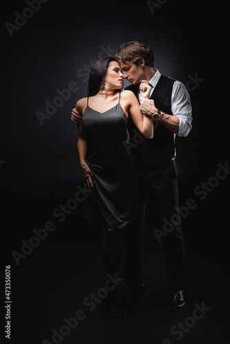 full length of man in vest and shirt hugging girlfriend in sexy slip dress on black.