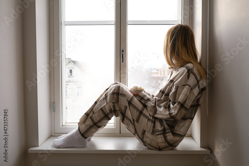 A girl in pajamas sits on the windowsill and holds a cup of cocoa with marshmallows. Cozy winter days at home. Quarantine time