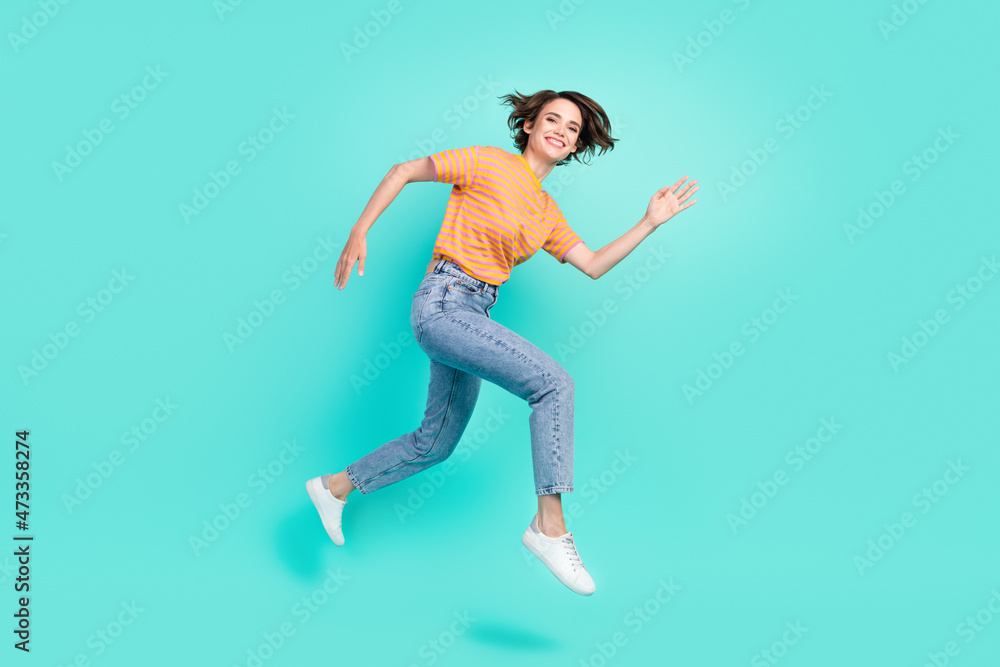 Photo of impressed cute young lady wear orange t-shirt jumping high running fast smiling isolated turquoise color background