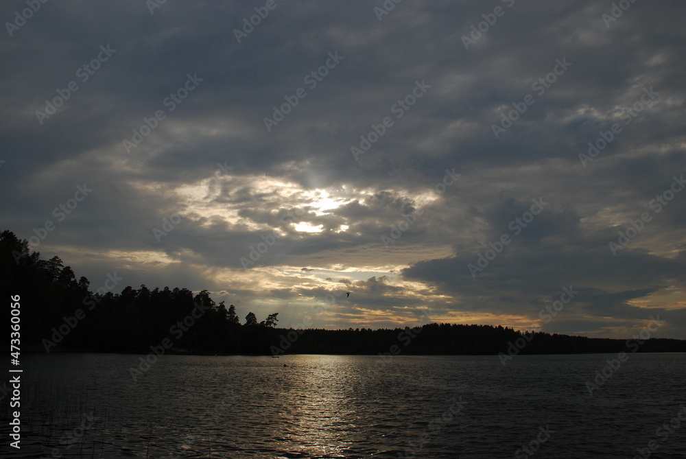 Summer cloudy day on the lake. There are small waves on the surface of the water, a dark forest can be seen in the distance. Gray-white clouds over the water, through which the sun shines through.