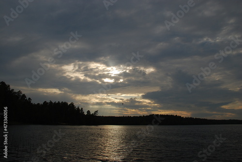 Summer cloudy day on the lake. There are small waves on the surface of the water, a dark forest can be seen in the distance. Gray-white clouds over the water, through which the sun shines through.