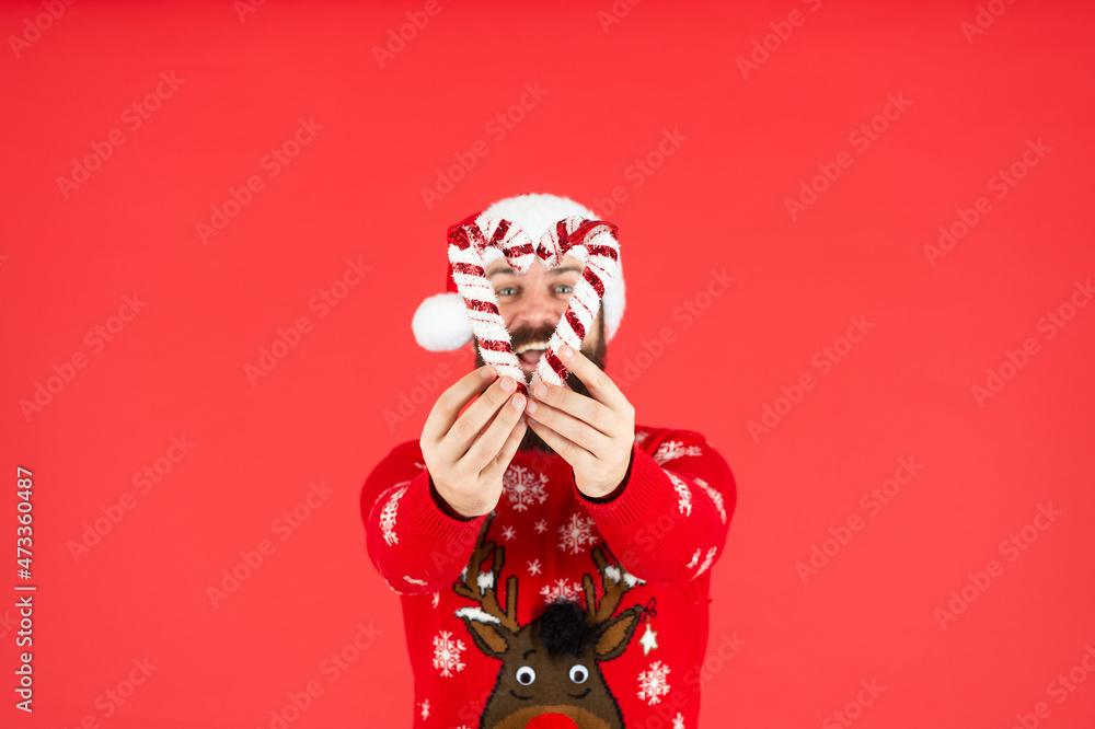 funny bearded guy in santa claus hat and sweater hold candy stick on red background, christmas