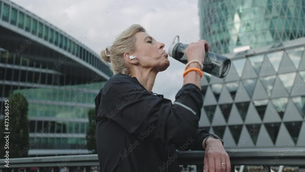 Mature woman in earphones drinking water after urban jogging Mature woman in earphones drinking water after urban jogging