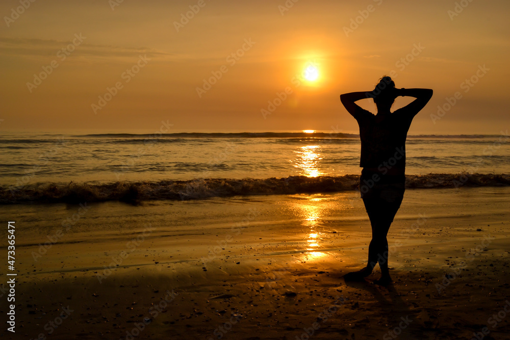 Silhouette of woman with arms on her head on the shore of a beach at sunset.