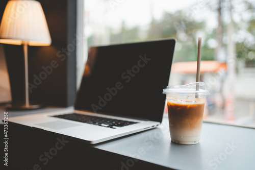 A glass of iced coffee on the table with blurred laptop background in cafe.