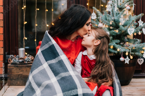 Mother hugging and kissing daughter wrapped in a blanket on Christmas tree background. Xmas mood. Single solo parenting holidays. Family time. Close family relationship. Selective focus.