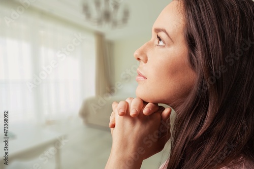 Religious young woman with clasped hands praying indoors at home