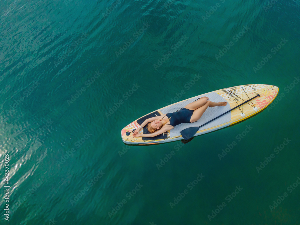 Fototapeta premium A woman lies on a sup, view from a drone. Young women Having Fun Stand Up Paddling in the sea. SUP. Red hair girl Training on Paddle Board near the rocks