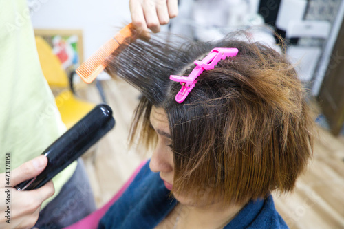 Transgender woman combing her hair with hairdryer, comb and hair straightener in the beauty salon by the professional stylist. Concept of beauty and hairdressing. Beauty and health center.