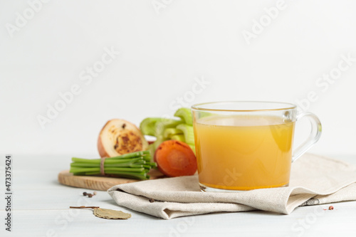 Vegetable or meat broth in a glass mug. Close-up, vegetables and roots in the background.