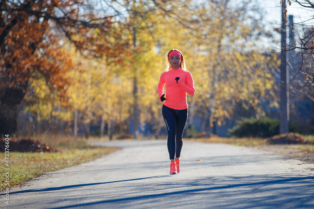 Obraz premium Young fitness woman running on the road in the cold morning in winter. Sporty girl in orange long-sleeve jogging in the sunny morning