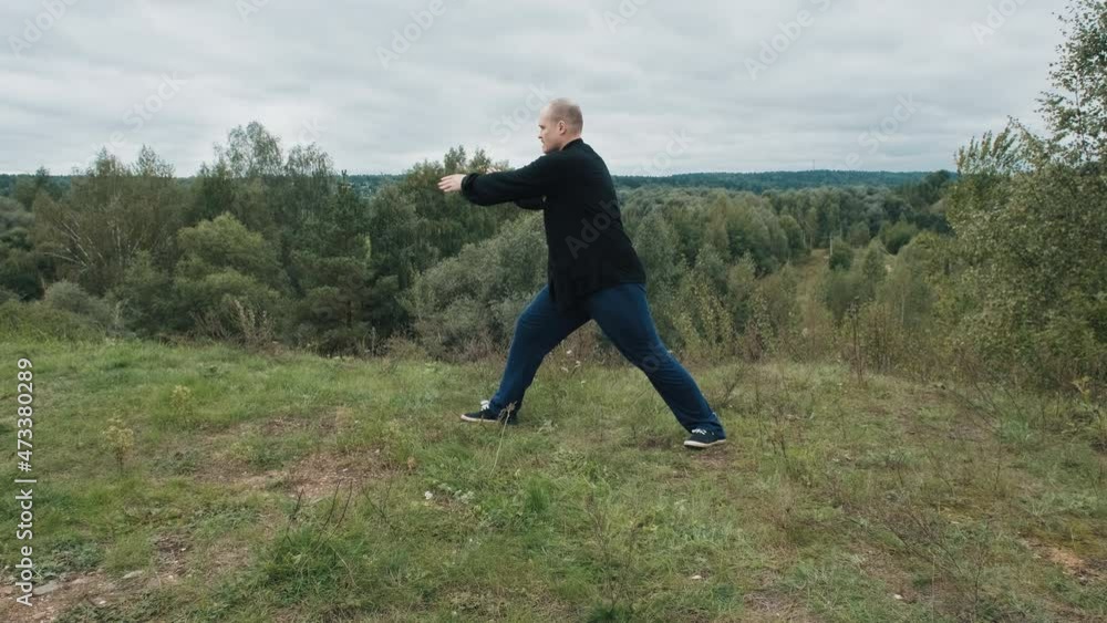 Middle aged Caucasian man practices wushu in the summer in a park on a hill. Traditional Chinese gymnastics qigong and Tai chi. Meditation and relaxation in nature. Healthy lifestyle and sports Middle aged Caucasian man practices wushu in the summer in a park on a hill. Traditional Chinese gymnastics qigong and Tai chi. Meditation and relaxation in nature. Healthy lifestyle and sports
