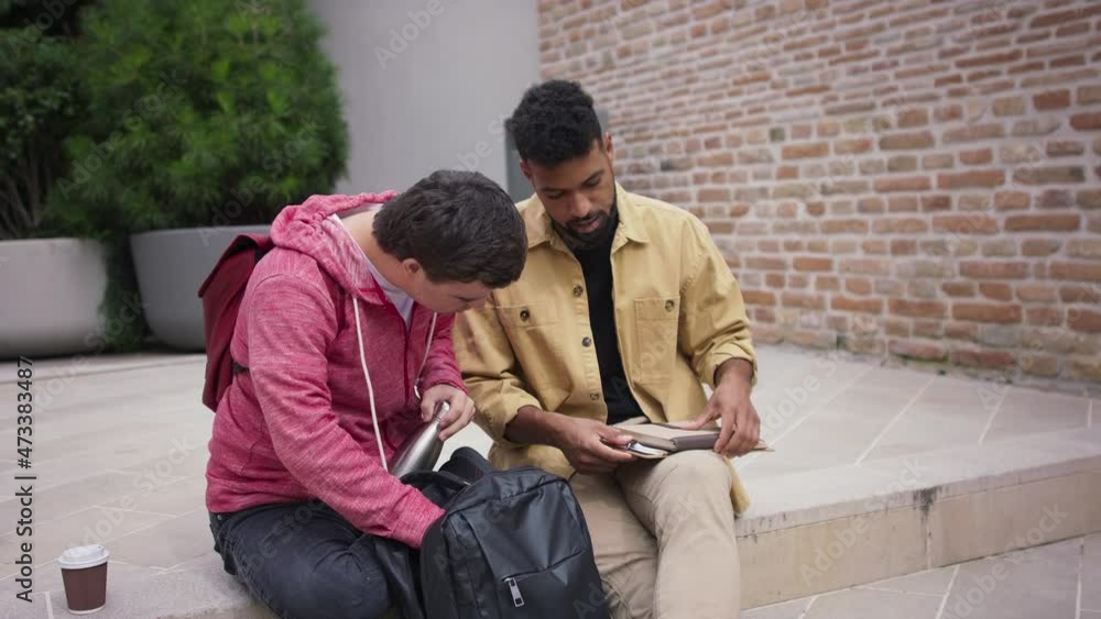 Happy young man with Down syndrome and mentoring friend sitting and talking on stairs outdoors in campus area. Happy young man with Down syndrome and mentoring friend sitting and talking on stairs outdoors in campus area.