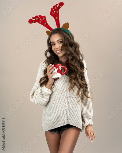 A beautiful young girl with long curly brown hair, in a sweater, poses on a light meringue background, holding a Christmas mug. Woman with reindeer horns on her head.