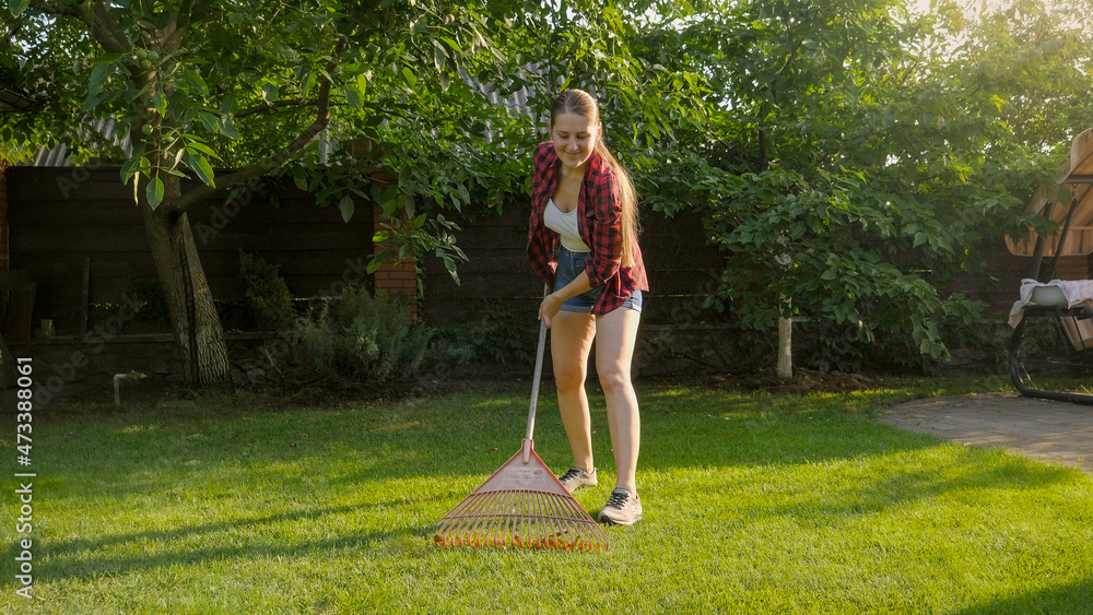 Young smiling woman working at house backyard and collecting fallen leaves with garden rake. Concept of houseworking, gardening and suburban life
