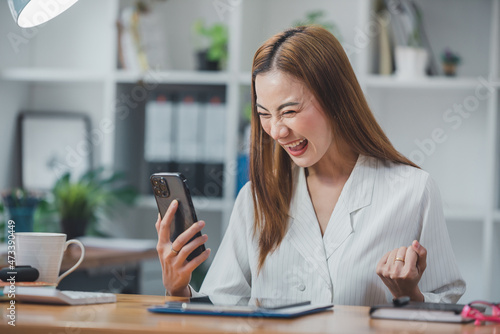 Excited Asian woman sitting at her desk Feel satisfied with the business going well. Very happy to receive letters from laptops and smartphones promoted at work. © Daenin