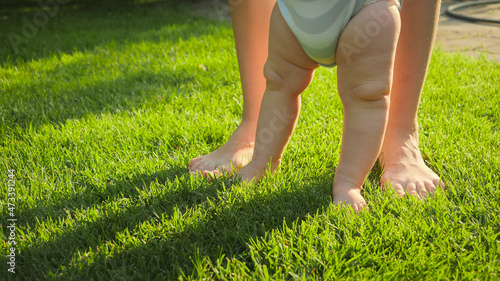 Closeup of mother supporting her baby son making first steps on fresh green grass lawn. Concept of healthy lifestyle, child development and parenting.