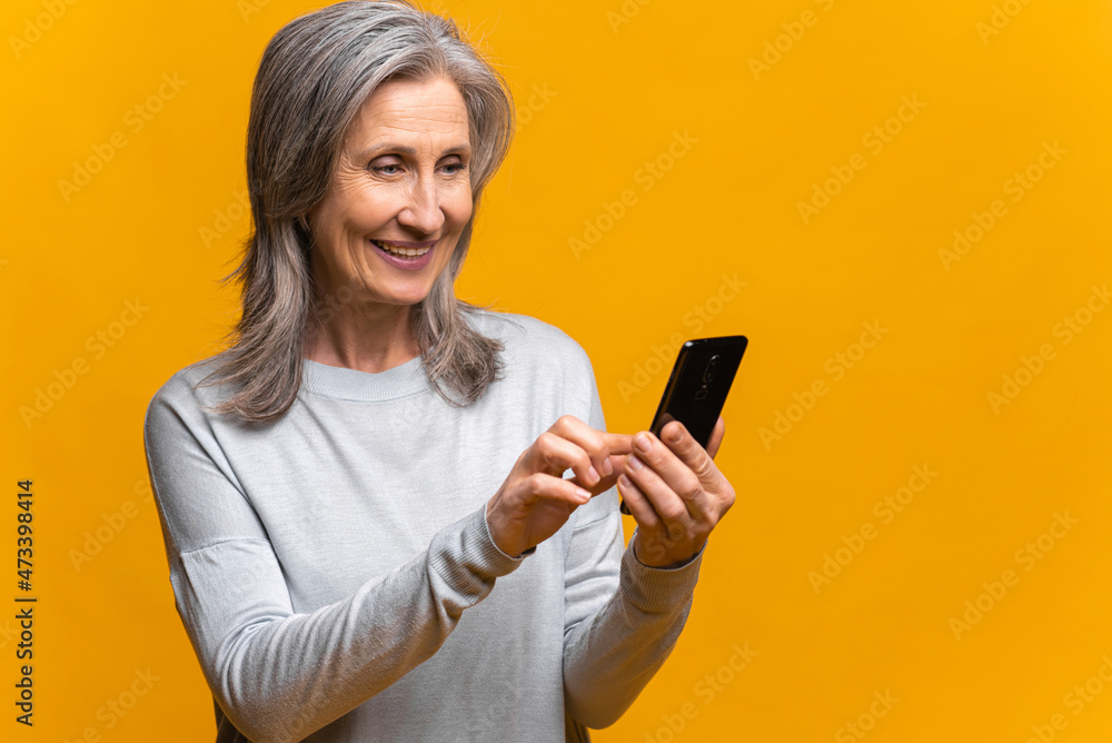 Smiling senior woman holding and using smartphone, carefree mature modern gray-haired lady enjoying online chatting in social media, using mobile app, isolated over yellow background Smiling senior woman holding and using smartphone, carefree mature modern gray-haired lady enjoying online chatting in social media, using mobile app, isolated over yellow background