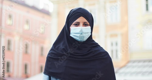 Attractive veiled female muslim standing on street and looking to camera. Adult arab woman in medical protective mask wearing hijab outdoors. Religin, people, culture concept. photo