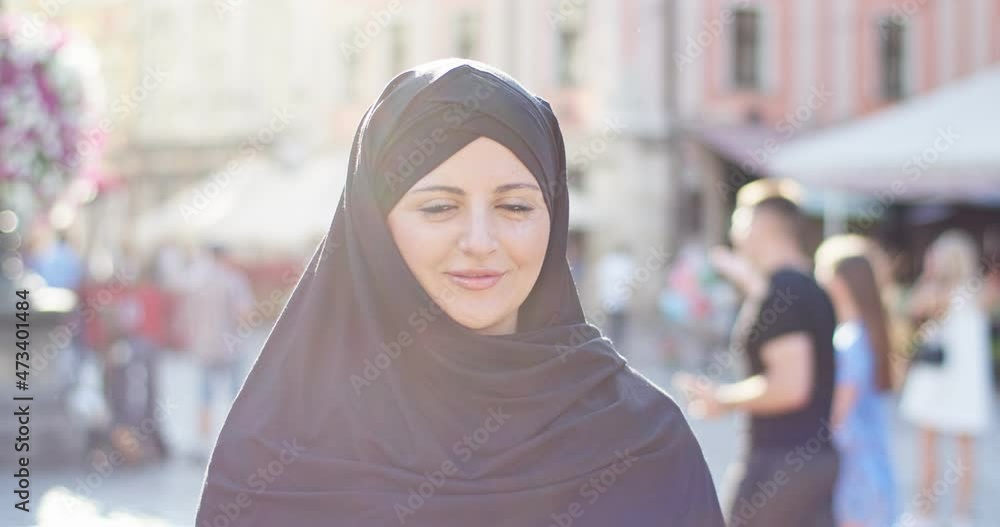 Portrait of good-looking veiled arab woman walking on street of city on sunny day. Young female muslim wearing black hijab, covering head, looking to camera and smiling. Religion, rights concept. Portrait of good-looking veiled arab woman walking on street of city on sunny day. Young female muslim wearing black hijab, covering head, looking to camera and smiling. Religion, rights concept.