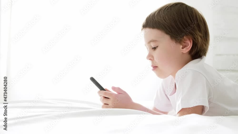 Happy 5 year boy play smartphone video game. Kid use mobile phone for gaming or learning. Smiling child in white shirt lie in home bedroom. Online education. Close-up. 4K video footage. Copy space. Happy 5 year boy play smartphone video game. Kid use mobile phone for gaming or learning. Smiling child in white shirt lie in home bedroom. Online education. Close-up. 4K video footage. Copy space.