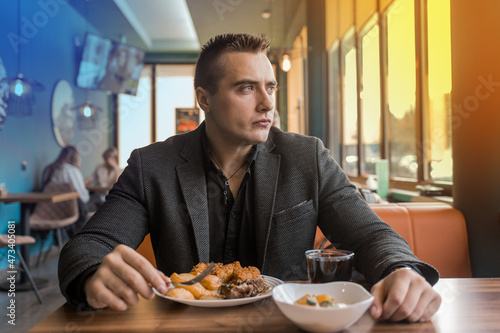 A serious  brooding stylish young man of European appearance  a businessman in a jacket sits at a table in a restaurant on a lunch break and looks out the window