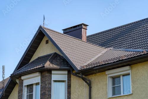 Corner of house with new brown metal tile roof and rain gutter. Metallic Guttering System, Guttering and Drainage Pipe Exterior