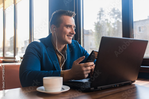 A business man stylish positive smiling businessman in an attractive European-looking suit works in a laptop, listens to music with headphones and sitting at a table in a cafe by the window