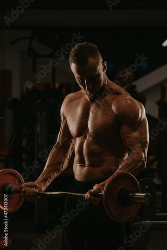 A bodybuilder with a beard is doing bicep curls with a short barbell in a gym. A photo of a torso of a naked muscular man during a workout.