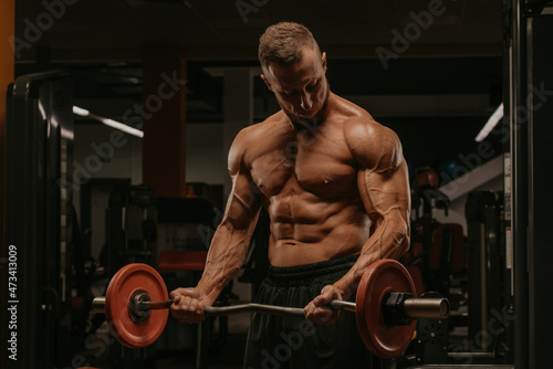 A bodybuilder with a beard is doing bicep curls with an EZ-barbell in a gym. A photo of a torso of a naked muscular man during a workout. © Roman Tyukin