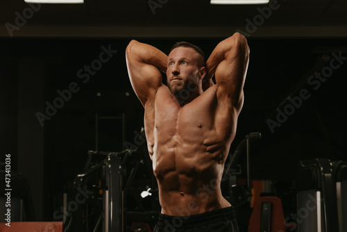 A bodybuilder with a beard is doing a stomach vacuum pose during a workout in a gym.