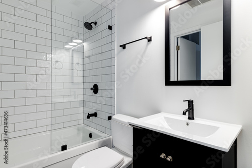 A small modern bathroom with a dark vanity, mirror frame, and hardware. White subway tiles line the bathtub and shower with black faucets. photo