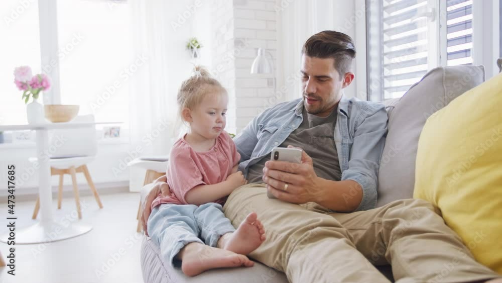 Young father sitting on sofa, holding his little daughter and using smartphone indoors at home Young father sitting on sofa, holding his little daughter and using smartphone indoors at home